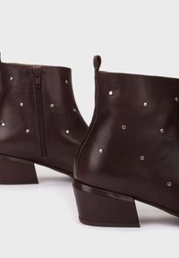 Bruine leren enkelboots met een ritsluiting, voorzien van zilveren studs aan de zijkanten en een lage, dikke hak. Gladde textuur.