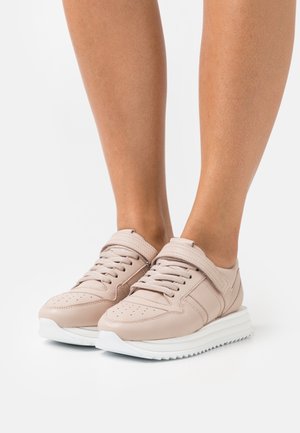 Sneaker beige chiaro con lacci e cinturino in velcro, indossate su gambe nude, che mostrano suole bianche spesse su uno sfondo bianco semplice.
