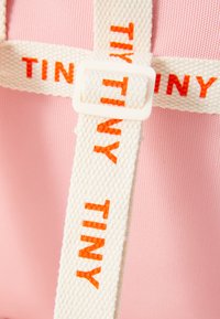 Rosa stoffveske med hvit vevet stropp som har "TINY" skrevet med store oransje bokstaver. Stropp sikret med en hvit plastspenne.