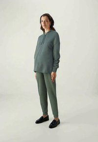 Pārlieku liels pelēkzaļš džemperis ar auklu, saskaņots ar atbilstošām šaurām biksēm, valkāts ar melnā krāsā apaviem uz neitrālā fona.