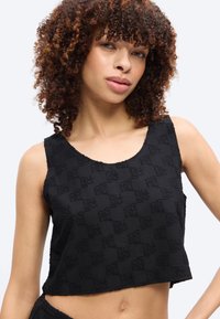 Γυναίκα με σγουρά μαλλιά, που φοράει μαύρο cropped τοπ χωρίς μανίκια με υφασμένο μοτίβο μονόγραμμα, ποζάροντας μπροστά σε απλό φόντο.