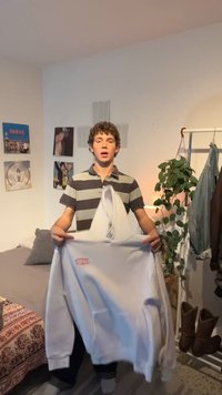 Ung person med krøllete hår holder opp en hvit hettegenser på et soverom med veggkunst, en plante og et klesstativ med jakker og støvler.