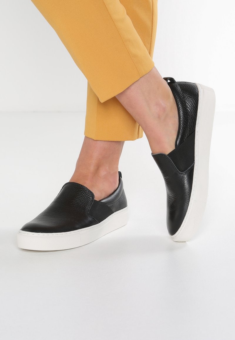 Baskets à enfiler en cuir noir avec semelles en caoutchouc blanc portées avec un pantalon court jaune moutarde, les jambes croisées.