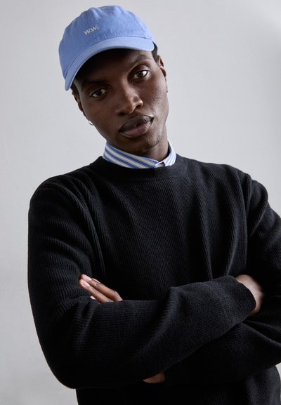 Zwart gestructureerd sweater over een lichtblauw gestreept shirt, afgewerkt met een lichtblauwe pet met witte letters "W.W.".