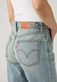 Άτομο που φοράει ανοιχτό μπλε τζιν Levi's με πίσω τσέπες και ορατό δερμάτινο ταμπελάκι Levi Strauss & Co στη ζώνη.