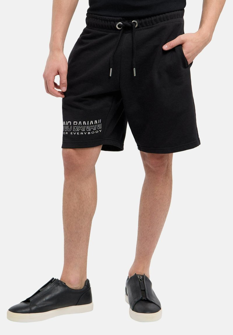 Man in zwarte trekband shorts met de tekst "MOO BANANA VOOR IEDEREEN" op het linkerpantbeen en zwarte instap sneakers met witte zolen.