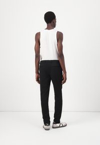 Sorte skreddersydde bukser, hvit ribbestrikket tank topp, grå-og-svarte sneakers. Modell står foran en nøytral bakgrunn og viser frempassformen og stilen på plagget.