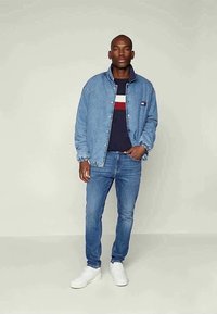 Denimjakke med frynser, blå langærmet skjorte med røde og hvide striber, lyseblå jeans og hvide sneakers.