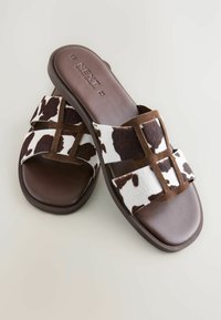 Braune Slide-Sandalen mit Kuhfell-Muster über den Riemen, einer glatten braunen Sohle und samtartigen Akzenten für zusätzliche Textur.