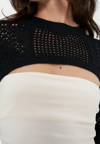 Gros plan d'une femme portant un pull court noir en maille ajourée sur un top blanc sans bretelles, montrant son cou et le haut de sa poitrine.