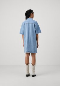 Lys blå denimkjole med korte ermer, lys søm og en avslappet passform, sammen med hvite cowboy-støvler og en liten tatovering synlig.