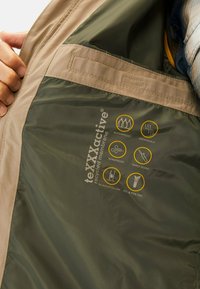 Groene waterdichte jas met een binnenkant in tan, uitgerust met een gerecycleerde membraan en pictogrammen voor waterdichtheid, ademend vermogen en winddichtheid.