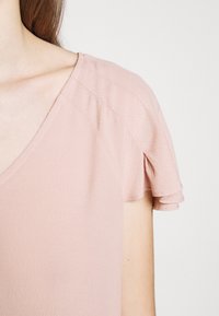 Jasnoróżowa bluzka z krótkim rękawem z dekoltem w kształcie litery V i subtelnym falbaną na rękawach, wykonana z teksturowanej, miękkiej tkaniny.