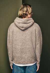 Beige strikket hættetrøje med en blød tekstur, der har en dyb hætte og en afslappet pasform, parret med blå jeans. Bagudsigten mod en mørk baggrund.