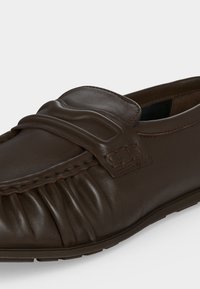 Καφέ δερμάτινο μοκασίνι με πιέτες στη μύτη και διακοσμητικό λουράκι στο πάνω μέρος του παπουτσιού.