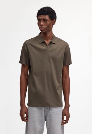 Giovane uomo che indossa una polo a maniche corte verde oliva e jeans grigio chiaro, in piedi contro uno sfondo bianco, guarda a sinistra.