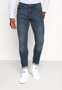 Blue slim-fit jeans with faded areas, five pockets, and a button closure. Paired with rolled cuffs and white sneakers.