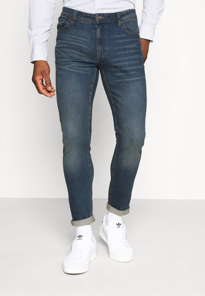 Blue slim-fit jeans with faded areas, five pockets, and a button closure. Paired with rolled cuffs and white sneakers.