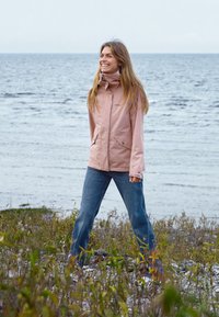 Lichtroze waterdichte jas met hoge kraag, met voorzakken en ritssluiting, gecombineerd met blauwe jeans voor een zeeachtergrond.