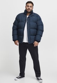 Navy puffer jacket with a high collar, button closure, quilted texture, paired with black pants and black sneakers.
