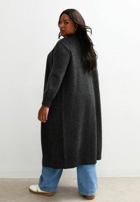 Gros cardigan gris foncé en maille douce avec une coupe décontractée, doté de manches longues et de poches latérales, associé à un jean bleu clair.
