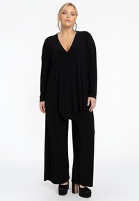 Zwarte vloeistof jumpsuit met lange mouwen en een diepe v-hals. Kenmerken zijn wijde pijpen en een gelaagde uitstraling, gecombineerd met platformschoenen.