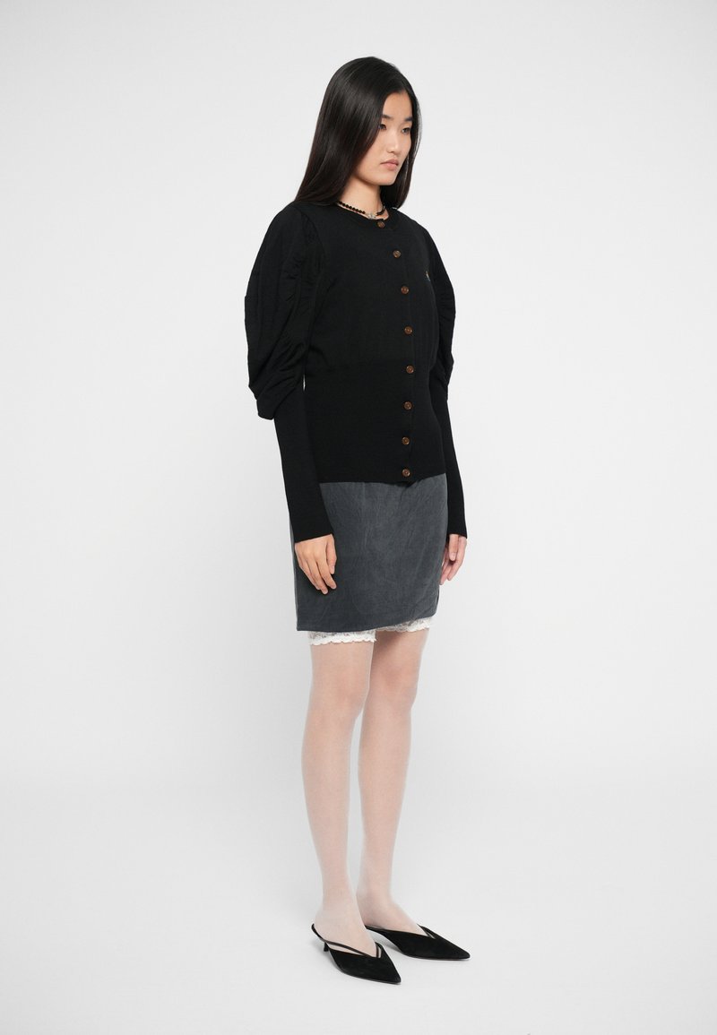 Cardigan nero con maniche a palloncino e bottoni in legno, abbinato a una mini gonna grigia con bordo in pizzo, completato con mule nere.
