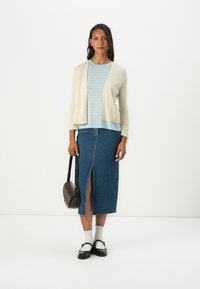 Un cardigan beige clair porté sur un t-shirt rayé bleu et blanc, associé à une longue jupe en denim avec une fente devant, des chaussures noires et un sac marron.