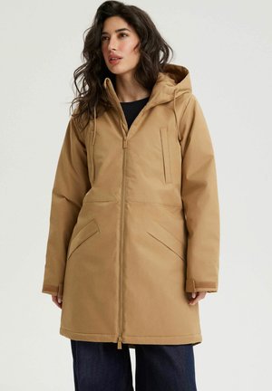 Kobieta w beżowej zimowej kurtce z kapturem, z przodu z zamkiem błyskawicznym i ukośnymi kieszeniami, w ciemnych spodniach, stojąca na jednolitym jasnym tle.