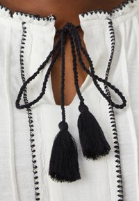 Bílá blůza s černým třásňovým uzlem na výstřihu; má texturovanou látku s černým prošíváním podél okrajů a límce.