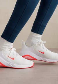Baskets blanches avec des accents rouges, présentant un dessus texturé et des semelles rembourrées, portées avec des chaussettes blanches mi-mollet et des leggings bleu marine.