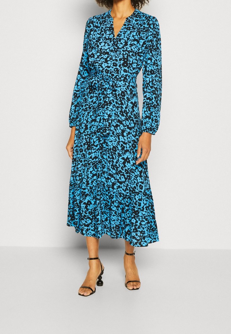 Femme portant une robe midi à manches longues imprimée fleurs bleu et noir avec un décolleté en V et des talons noirs à bride cheville sur fond blanc.