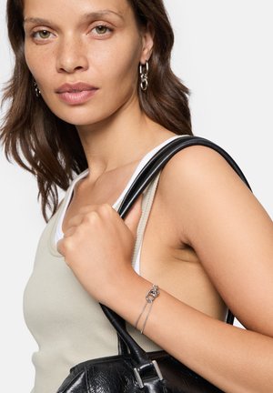 Silver bracelet with a knot detail and chain, worn on a bare wrist. The model holds a black handbag with smooth texture, wearing a beige top.