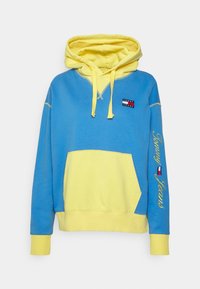 Sudadera color bloque en azul y amarillo con un bolsillo delantero, capucha con cordón y "Tommy Jeans" bordado en la manga. Material de algodón.