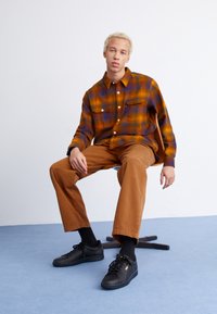 Brown plaid shirt with orange and blue tones, two chest pockets; tan pants; black sneakers; seated pose on a chair against a light background.