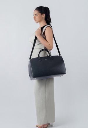 Ženska v bež obleki brez rokavov, ki nosi veliko črno usnjeno torbo za na ramo, na sivi podlagi.