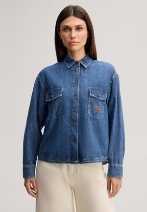Donna con capelli castani lisci che indossa una camicia di jeans blu con tasche sul petto e pantaloni beige su uno sfondo uniforme.
