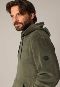 Olijfgroene hoodie van zacht, gestructureerd materiaal met een capuchon met trekkoord, zwarte accenten en een ronde logo patch op de mouw.