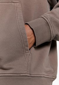 Brown sweatshirt featuring a ribbed cuff and side pocket. The fabric has a smooth texture with stitching detail around the edges.
