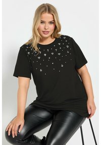 Camiseta negra de manga corta con decoración de piedras de strass dispersas en el frente. Tejido suave, corte relajado, combinada con leggings negros brillantes.