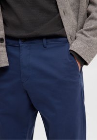 Tmavě modré kalhoty chino s hladkou texturou, opatřené zipem a knoflíkovým zapínáním, a postranními kapsami. Nošeny s šedým pleteným svetrem.