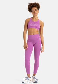 Vaaleanpunainen urheiluliivi ja leggingsit joustavasta kankaasta, joissa on logo liivissä ja hienovarainen muotoilu leggingsissa. Yhdistetty urheilukenkiin.
