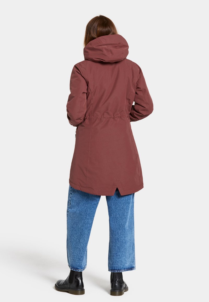 Lång vinröd jacka med dragsnören i midjan, stor huva och ryggslits. Bärs över lösa blå jeans och svarta ankelstövlar. Slen yta.