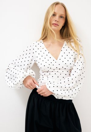 Mladá žena s dlhými blond vlasmi, oblečená v bielej blúzke s čiernymi bodkami a dlhými rukávmi, upravujúca manžetu, viditeľná čierna sukňa.