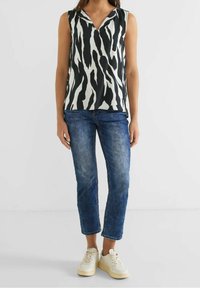 Žena v černobílém topu se zebřími pruhy bez rukávů, modrých zkrácených džínách a bílých teniskách, stojící proti jednoduchému bílému pozadí.