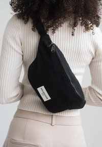 Black canvas sling bag with a zip closure and white tag featuring text. It has a unique triangular shape and a sturdy strap.