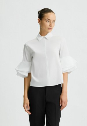 Chemise blanche à boutons avec un col et des manches trois-quarts à volants superposés. Fabriquée dans un tissu léger et lisse.