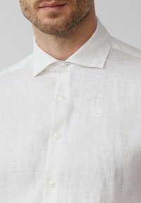 Weißes Leinen-Hemd mit Knopfleiste, das einen spitzen Kragen und fünf Knöpfe aufweist und einen strukturierten Stoff mit dezenten, natürlichen Fasern zeigt.