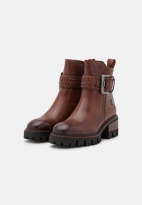 Hnědé kožené kotníkové boty mají pletený pásek s přezkou, texturovaný podpatek a robustní černou podrážku. Hladký povrch s dekorativními akcenty.