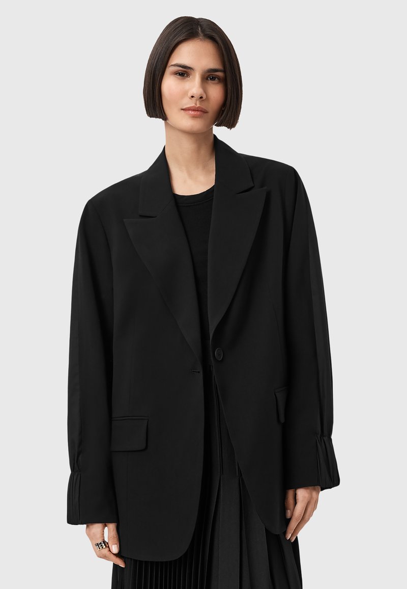 Blazer oversize nero realizzato in tessuto liscio, con rever a punta, chiusura a un bottone e due tasche frontali. Dettaglio delle maniche plissettate.